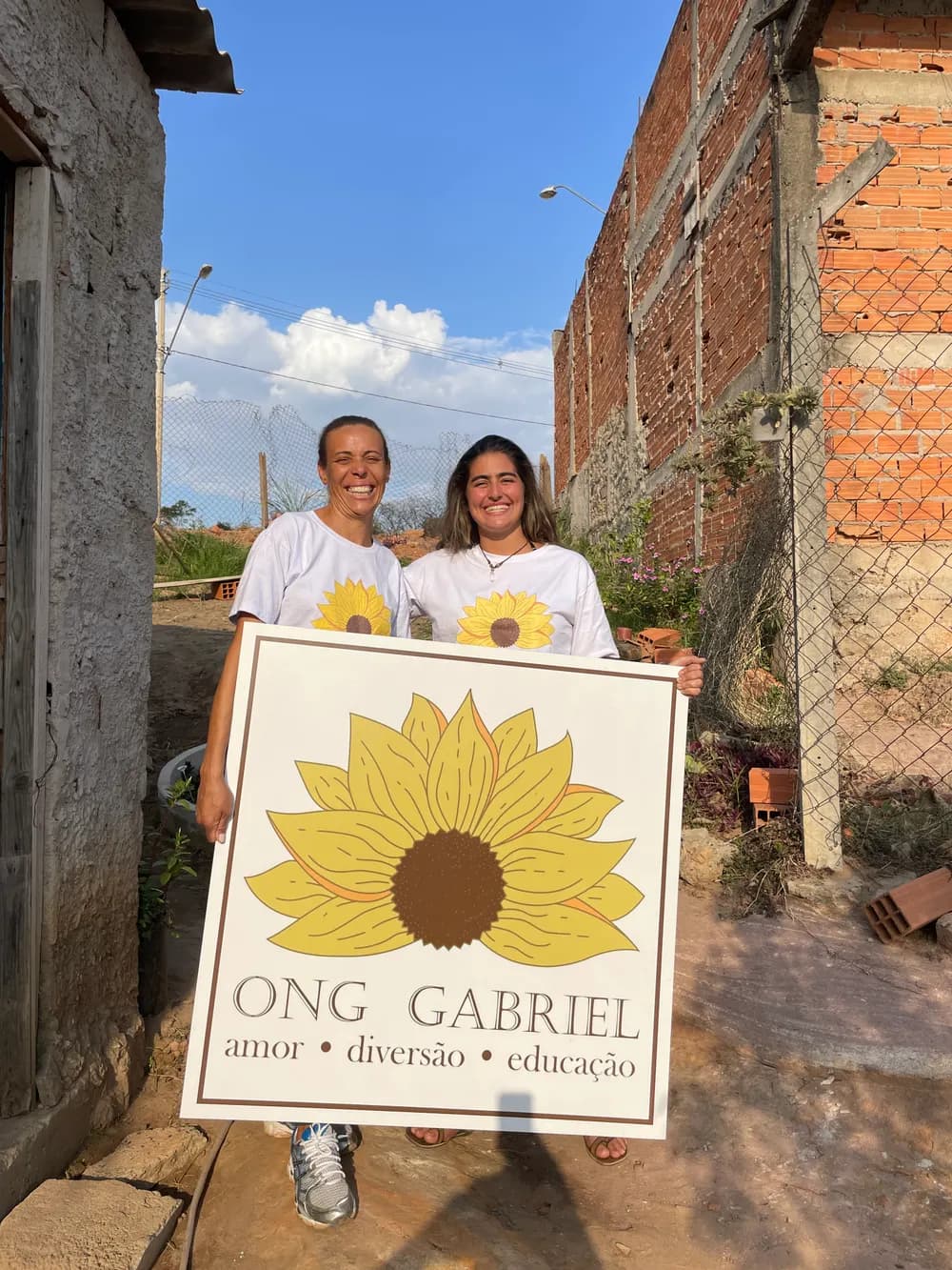 Nádia e Clara sorriem segurando o cartaz da ONG Gabriel, com um girassol e as palavras amor, diversão e educação.