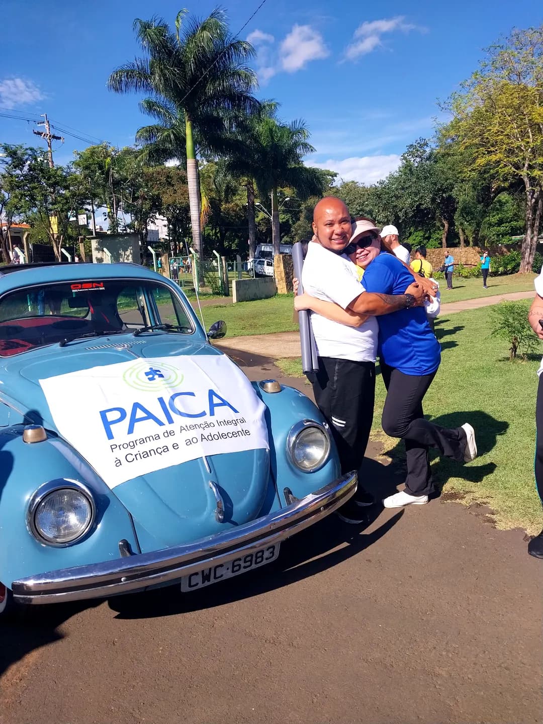 Duas pessoas se abraçam ao lado de um Fusca azul com a faixa do PAICA, em um parque.