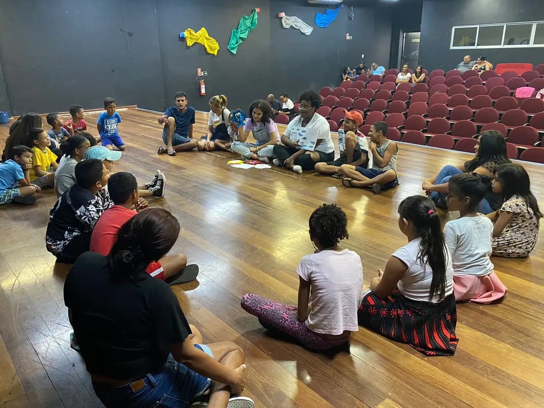 Crianças e jovens sentados no chão de um auditório durante uma atividade.