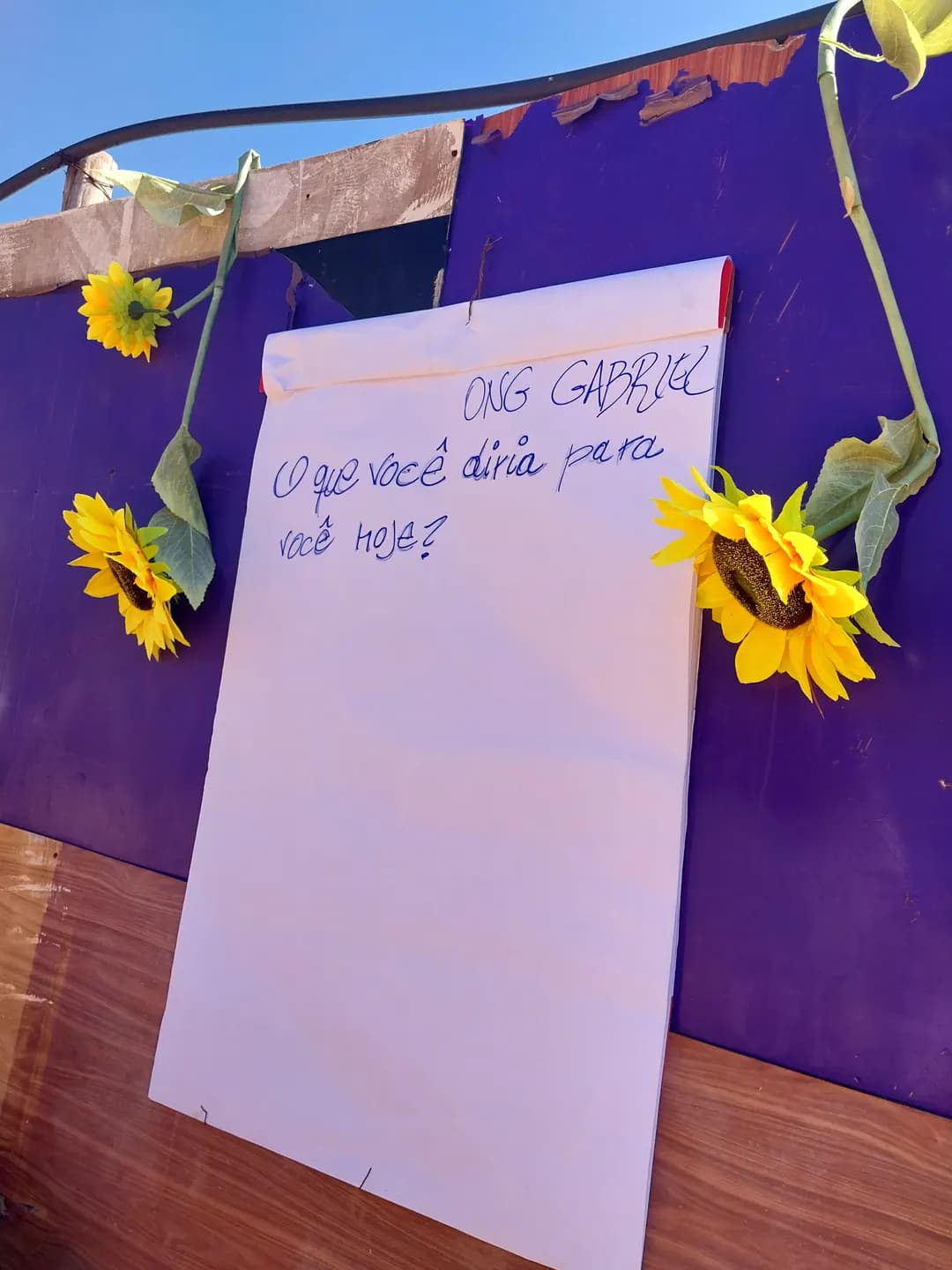 Cartaz da ONG Gabriel com a pergunta “O que você diria para você hoje?”, preso a um painel roxo com girassóis.