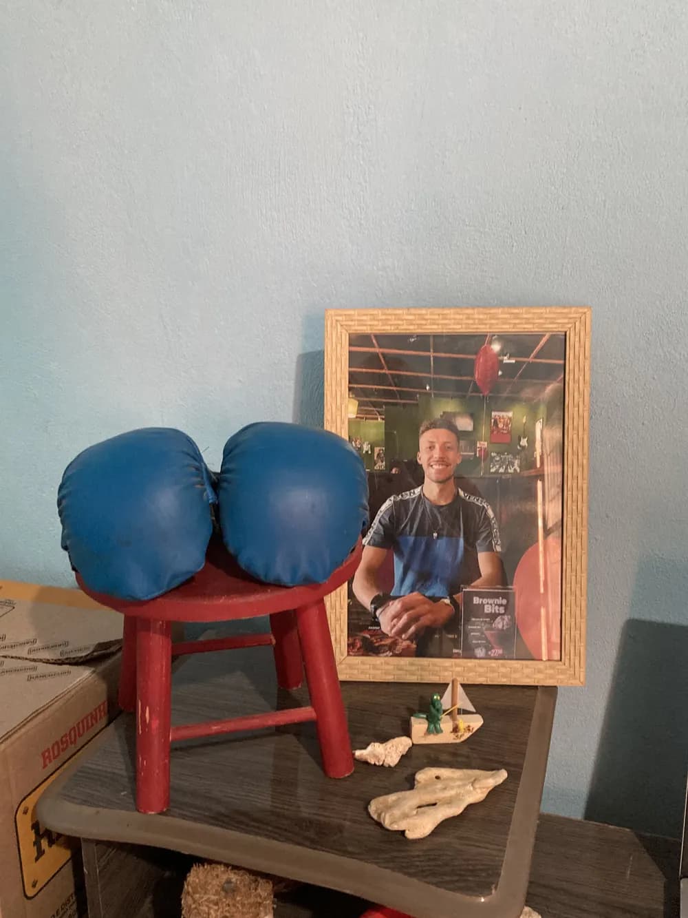 Um par de luvas de boxe azuis sobre um banquinho vermelho, ao lado de um porta-retrato com a foto de Gabriel sorrindo.